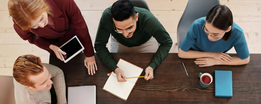Aus der Vogelperspektive sitzen 4 Menschen an einem braunen Tisch in einem Business Meeting. Alle lächeln und haben Spaß am Meeting.