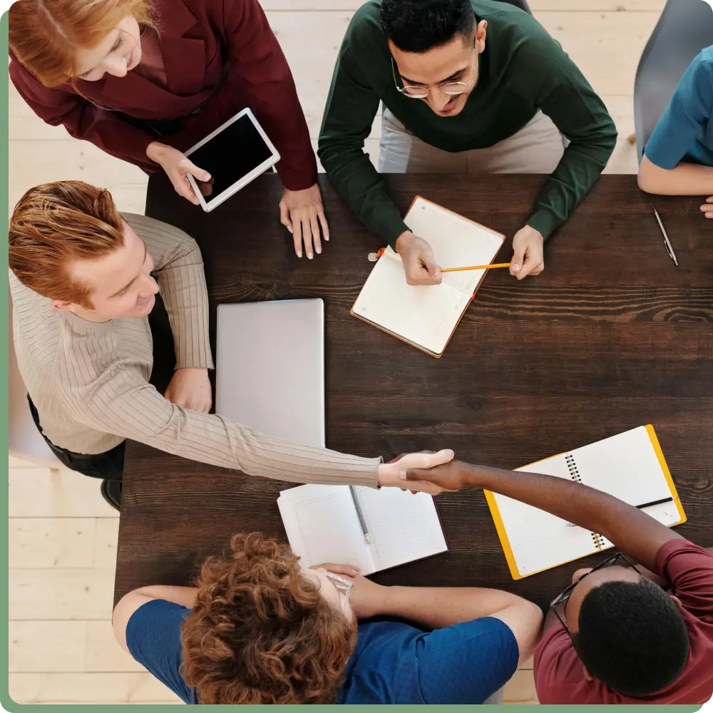 Aus der Vogelperspektive sitzen 6 Menschen an einem braunen Tisch. 2 Männer schütteln sich die Hände. 